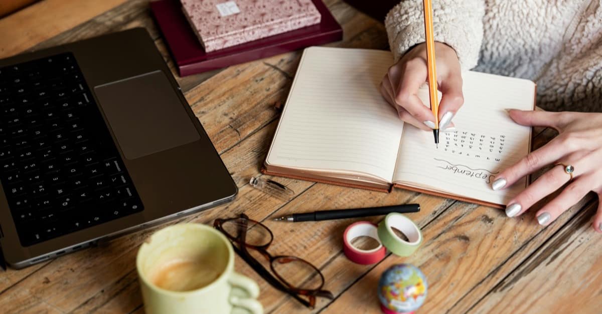 Overhead view of a wooden desk: a woman's hands writing a monthly plan in an open notebook, beside a laptop, a coffee mug, and reading glasses. The weekly planning habit at the center of burnout recovery.