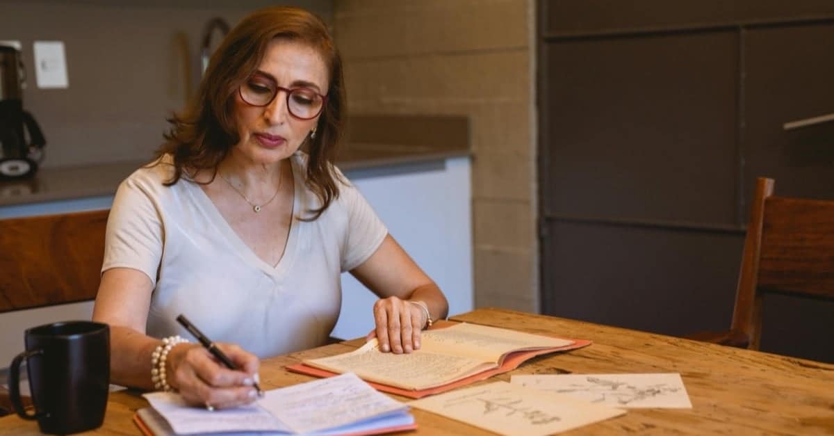 A woman in her early fifties sits at a kitchen table in soft light, calmly writing in a notebook beside an open book and coffee, a small daily self-discipline practice.